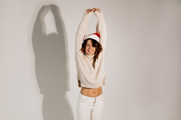 Wonderful pretty young lady with wavy hair dressed white sweater and jeans and santa hat posing with hands up 