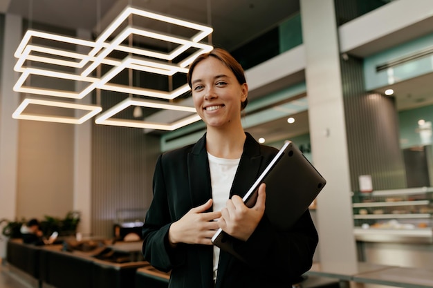 Wonderful stylish business woman in suit is holding laptop and smiling at camera