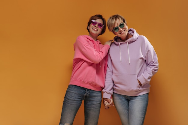 Замечательные стильные две женщины в прохладных солнцезащитных очках и розовых толстовках улыбаются и держатся за руки на оранжевом изолированном фоне.