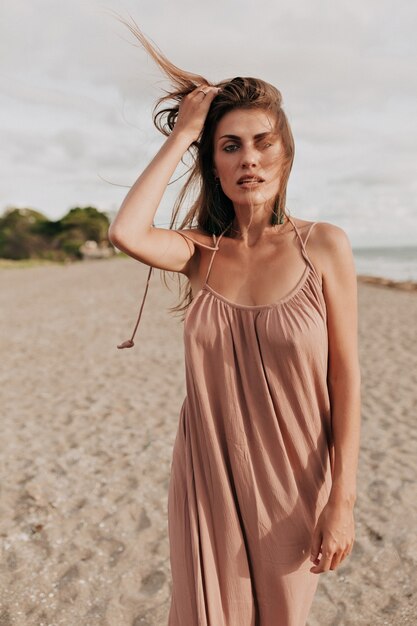 Wonderful stylish woman with long hair wearing long dress posing while walking on the beach