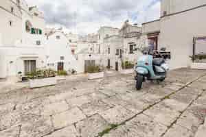 Foto gratuita splendida vista sulle strade vuote del centro storico martina franca con belle case dipinte di bianco in mezzo al verde. ciclomotore blu classico sullo sfondo di edifici antichi. bella giornata in un tour