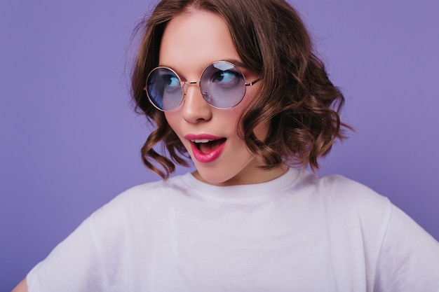 紫色の壁に優しく笑顔でポーズをとる流行の髪型の素晴らしい白人の女の子。インスピレーションを得た女性モデルはエレガントなサングラスをかけています。