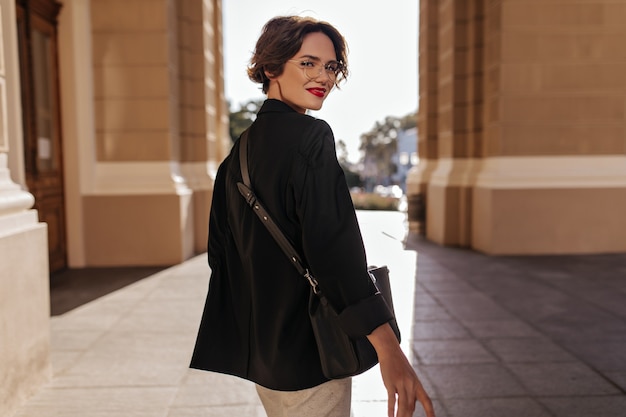 Wonderful woman in black jacket with dark handbag smiling at street. Short-haired woman in eyeglasses with red lips posing outside.