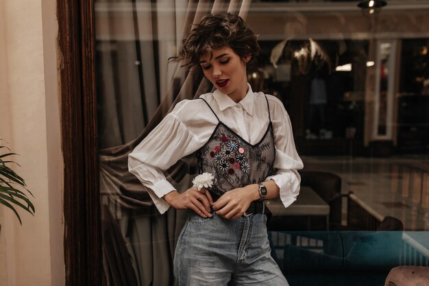 Wonderful woman in jeans and shirt with black lace and long sleeve posing in cafe. Short haired woman holding flower inside.