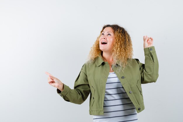 Wonderful woman pointing to the left side and back in green jacket, shirt and looking surprised. front view.