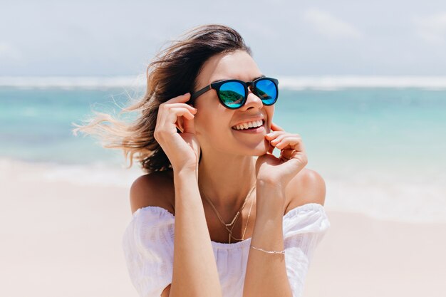 Wonderful woman in white attire and sparkle glasses posing with happy face expression in hot summer day. Pleasant caucasian woman standing near ocean on sky