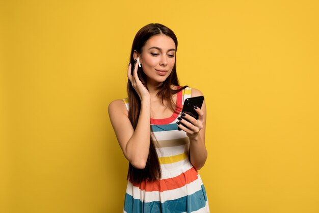 Wonderful woman with long hair and nude make up wearing bright dress listening music and holding smartphone