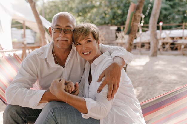 Wonderful woman with short blonde hairstyle in modern blouse smiling, sitting on hammock and hugging with her husband in eyeglasses on beach.