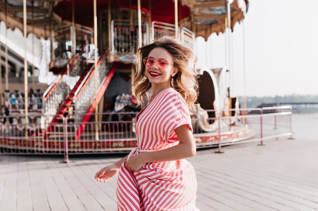 Wonderful young woman looking over shoulder while posing beside carousel. Laughing jocund girl in sunglasses expressing happiness in summer amusement park.