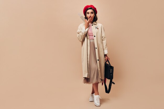 Wonderful young woman in stylish red beret and beige trench coat looks surprised and holds tickets on isolated background..