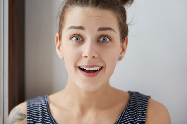 Wondering European female with beautiful blue eyes and open mouth . Astonishing facial expression of young brunette girl with tattoo on shoulder. Surprised emotions.