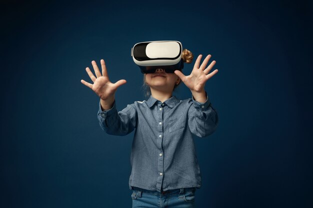 Wondering of feelings. Little girl or child in jeans and shirt with virtual reality headset glasses isolated on blue studio background. Concept of cutting edge technology, video games, innovation.