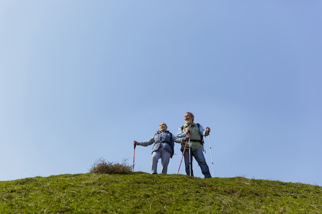 Удивлен и счастлив. Пожилая семейная пара мужчина и женщина в туристическом наряде гуляют по зеленой лужайке рядом с деревьями в солнечный день