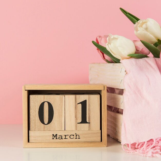 Wooden 1st march calendar near the crate with scarf and tulips against pink background