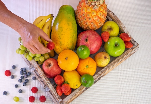 Wooden basket full of fresh fruit on a white background. citrus fruits, papaya, pineapple, banana, pear, apple, strawberry, pomegranate, blueberry. female hand holding a strawberry