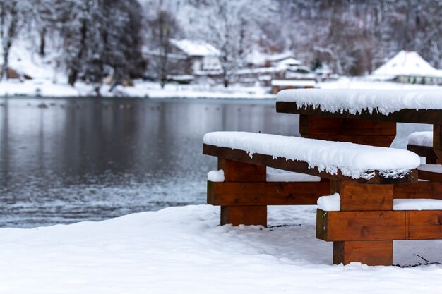 나무 벤치와 겨울 동안 눈에 덮여 나무로 둘러싸인 호수 근처 테이블
