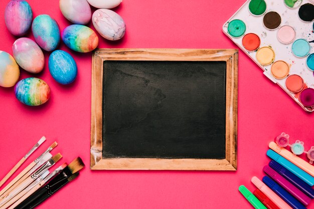 Wooden blackboard surrounded with easter eggs; paint brushes; felt tip pens and water color paint box on pink background