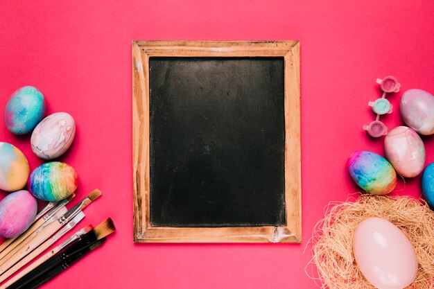 イースターエッグと木製の空白の黒板。ペイントブラシとピンク色の背景色