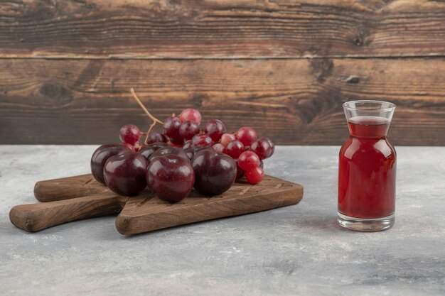 Wooden board of fresh red plums and grapes with glass of juice on marble surface. 