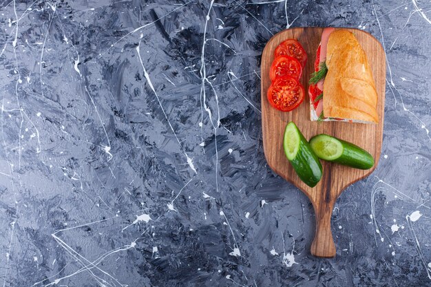 Wooden board of homemade sandwich and sliced vegetables on marble surface. 