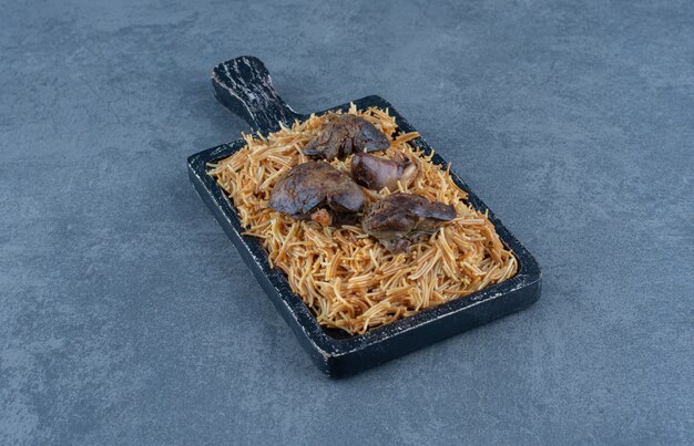 Wooden board of pasta with dry meat on stone table.