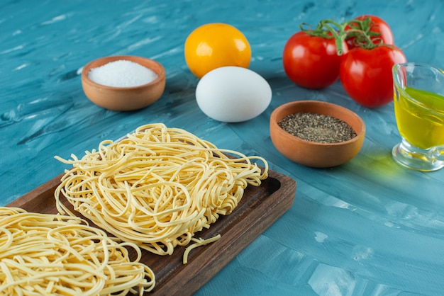 Free photo wooden board of raw noodles with condiments and tomatoes on blue surface