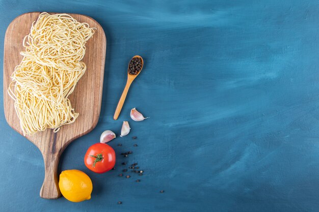 A wooden board of raw noodles with fresh red tomato and lemon on a blue background.. 