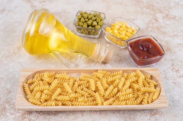 A wooden board of raw spiral macaroni and a glass bottle of oil