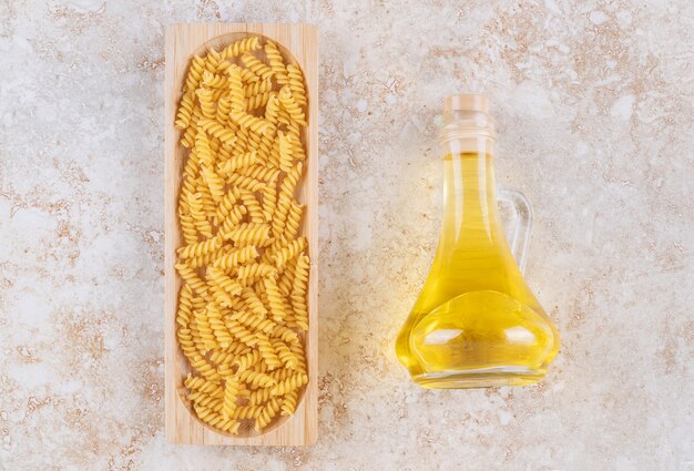 A wooden board of raw spiral macaroni and a glass bottle of oil