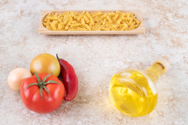 A wooden board of raw spiral macaroni and a glass bottle of oil