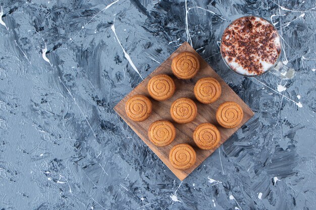 A wooden board of sweet round cookies with a glass cup of tasty hot coffee on a marble background. 