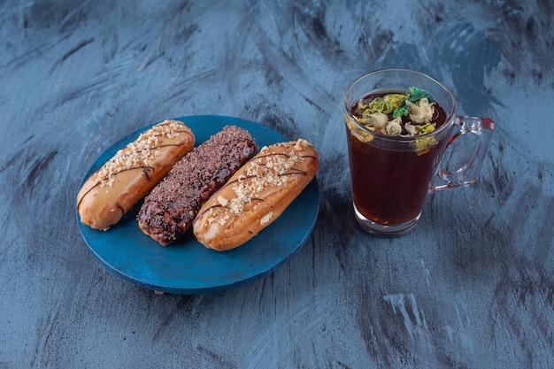 Wooden board with various sweet eclairs and cup of tea on marble surface.