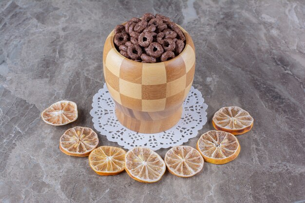 A wooden bowl of chocolate cereal rings with sliced dried orange fruit . 