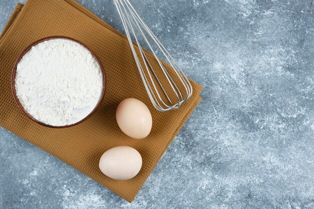 A wooden bowl of flour with two fresh chicken eggs and whisk.