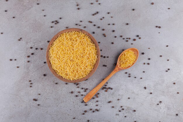 A wooden bowl full of raw dry Filini vermicelles with pepper corns .