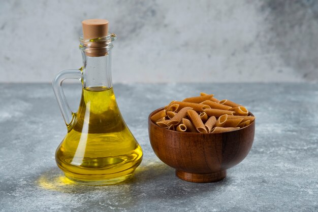 A wooden bowl full of raw grain pasta and a glass bottle of oil.