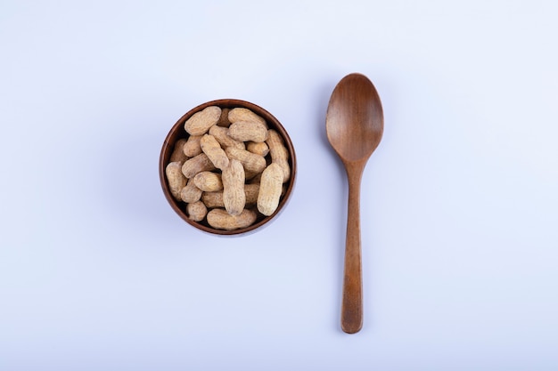 Wooden bowl full of unpeeled peanuts and spoon on white.