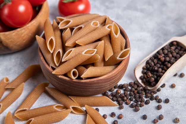 A wooden bowl of Italian uncooked dry pasta Penne rigate with tomatoes and pepper corns. High quality photo