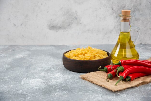 Wooden bowl of raw pasta with red chili peppers and oil on marble.