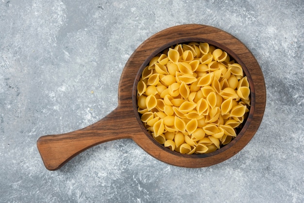 Wooden bowl of raw seashell pasta on cutting board.