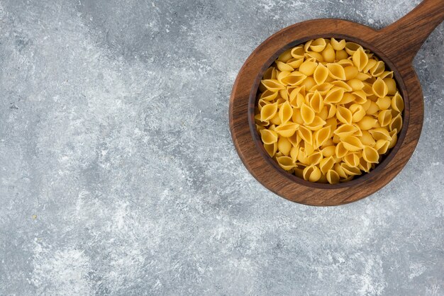 Wooden bowl of raw seashell pasta on cutting board.