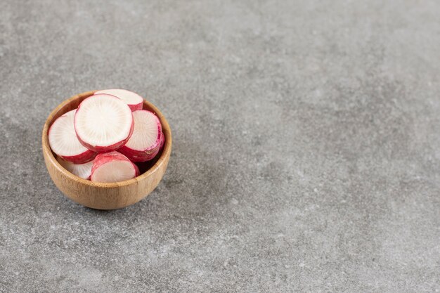 Wooden bowl of sliced red radish on marble surface. 