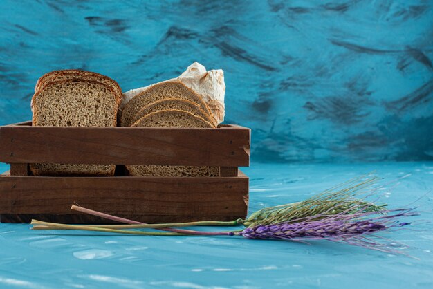 A wooden box full of sliced brown fresh bread with wheat ears on blue background . 