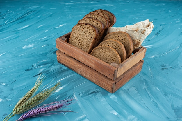 A wooden box full of sliced brown fresh bread with wheat ears on blue surface 