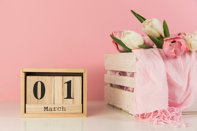 Wooden calendar showing 1st march near the crate with tulips and scarf against pink background