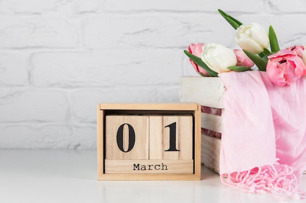 Wooden calendar with 1st march near the wooden crate with tulips and scarf on white desk