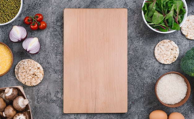 Wooden chopping board surrounded with vegetables and puffed rice cakes on concrete backdrop