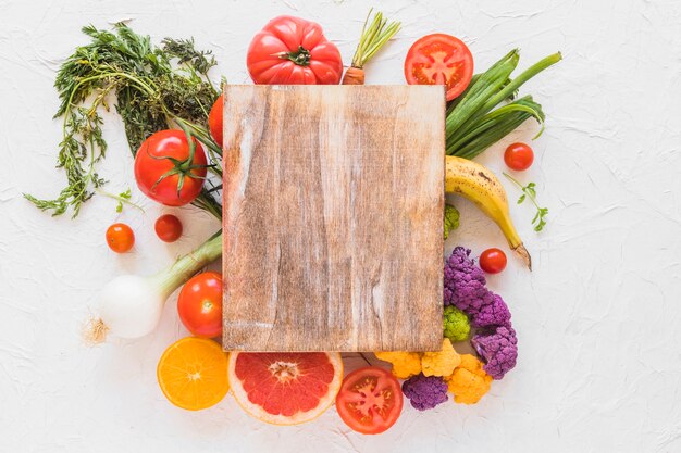 Wooden chopping board over the vegetables and fruits on white texture backdrop
