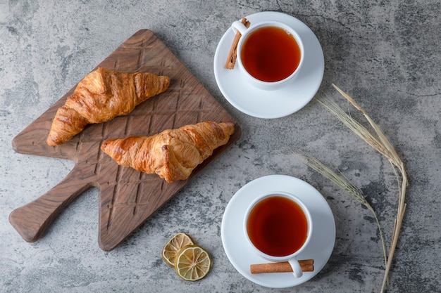 A wooden cutting board of fresh croissants and white cups of hot tea placed on a stone table. 