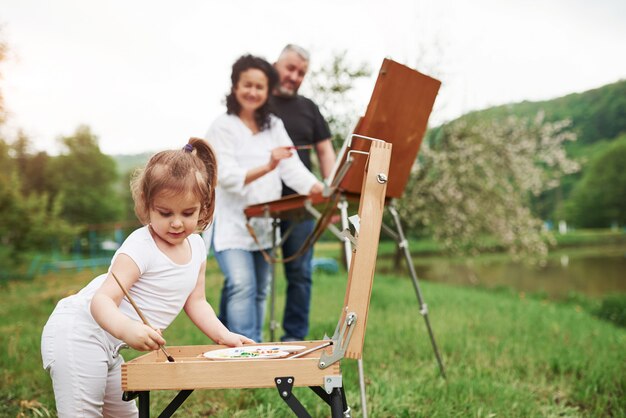 Wooden easel. Grandmother and grandfather have fun outdoors with granddaughter. Painting conception